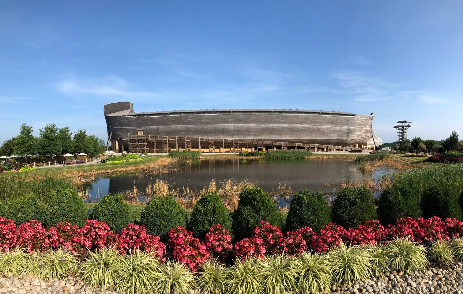 A large Ark replica is sitting next to a body of water surrounded by flowers.