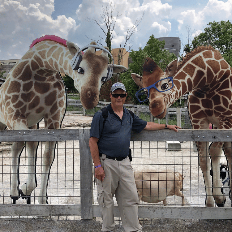 Man with giraffes wearing headgear and glasses at a zoo.