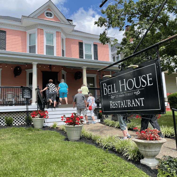 People enter Bell House Restaurant, a pink building with a porch, floral planters, and a black sign.