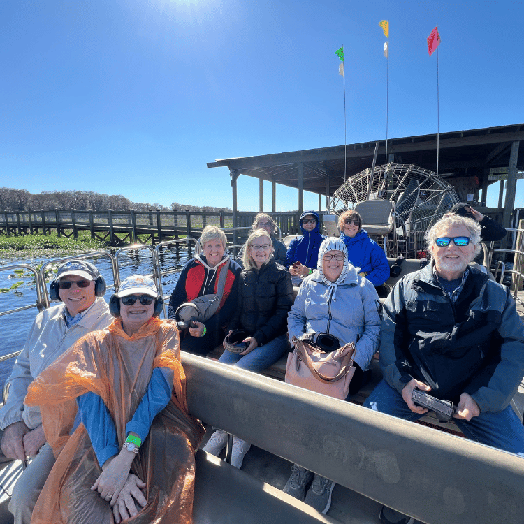 Group on airboat in a swamp; sunny day. People smiling and wearing coats and ear protection. Flags in the background.