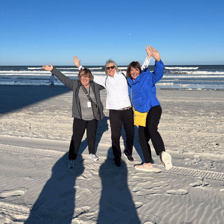 Three women with arms raised on a sunny beach, smiling.