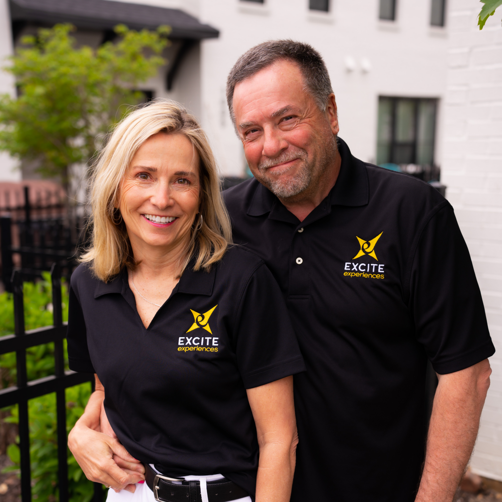 Couple in black polo shirts smiles outdoors; building in background.
