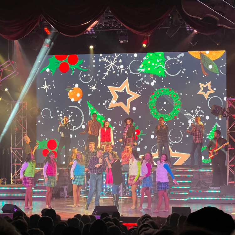 Musical performance with dancers in colorful outfits and holiday-themed backdrop. A crowd watches from the front.