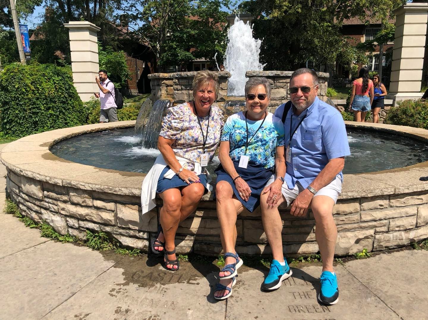 Three people are sitting on a stone wall in front of a fountain.