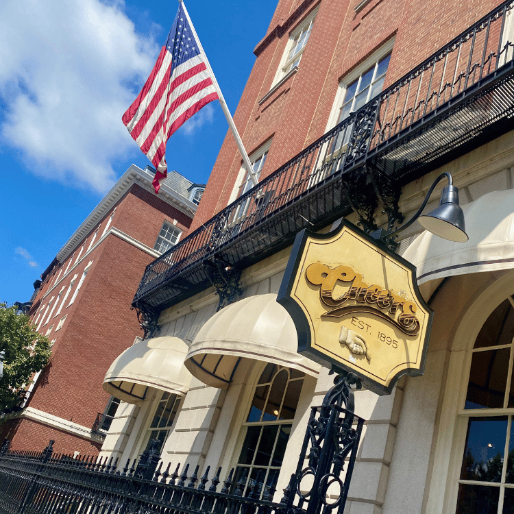 Exterior of Cheers bar in Boston with American flag. Red brick building, beige awnings, sign with logo.