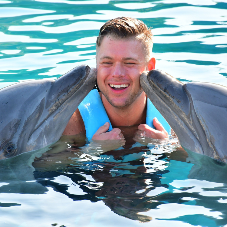 Man smiles as two dolphins touch his face in the blue water. He wears a blue life vest.