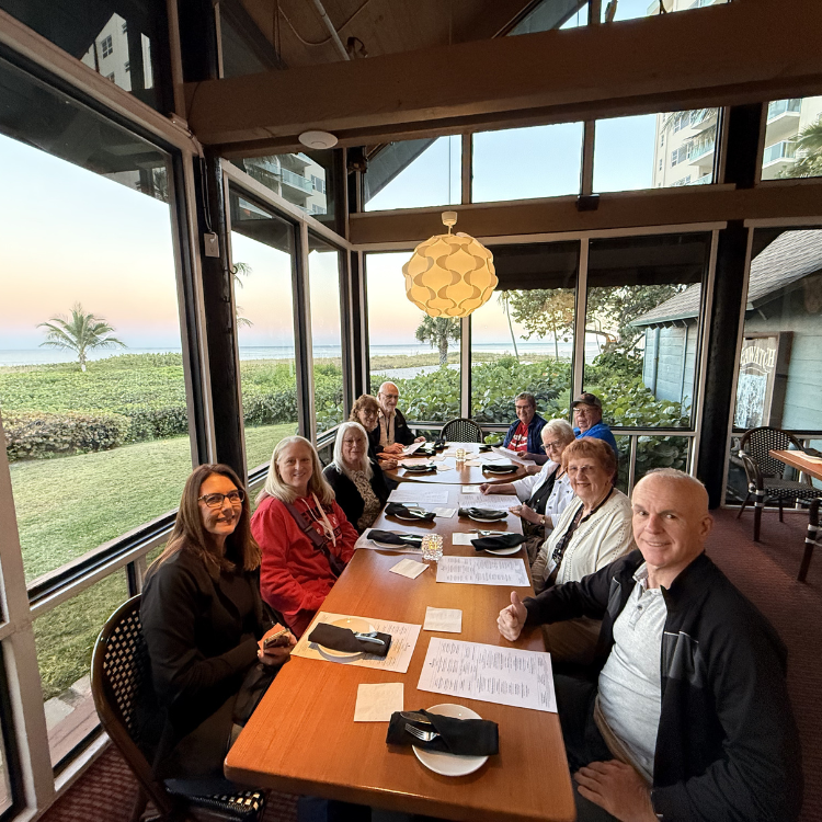 Group of people seated around a table in a restaurant with large windows overlooking a scenic ocean view.