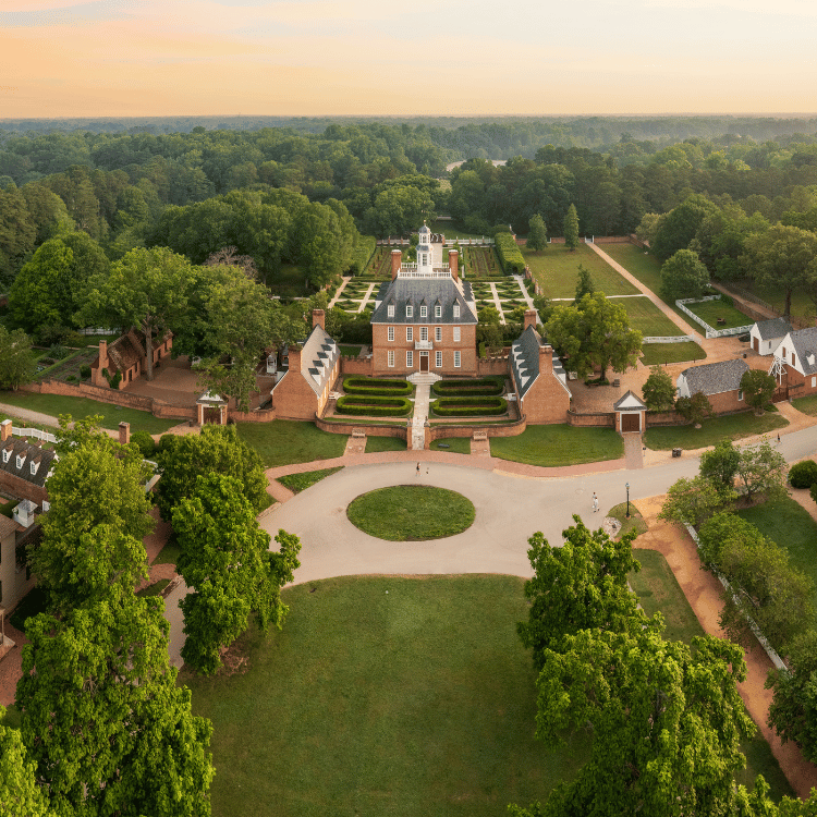 Aerial view of Colonial Williamsburg's Governor's Palace, surrounded by gardens and trees, bathed in golden light.