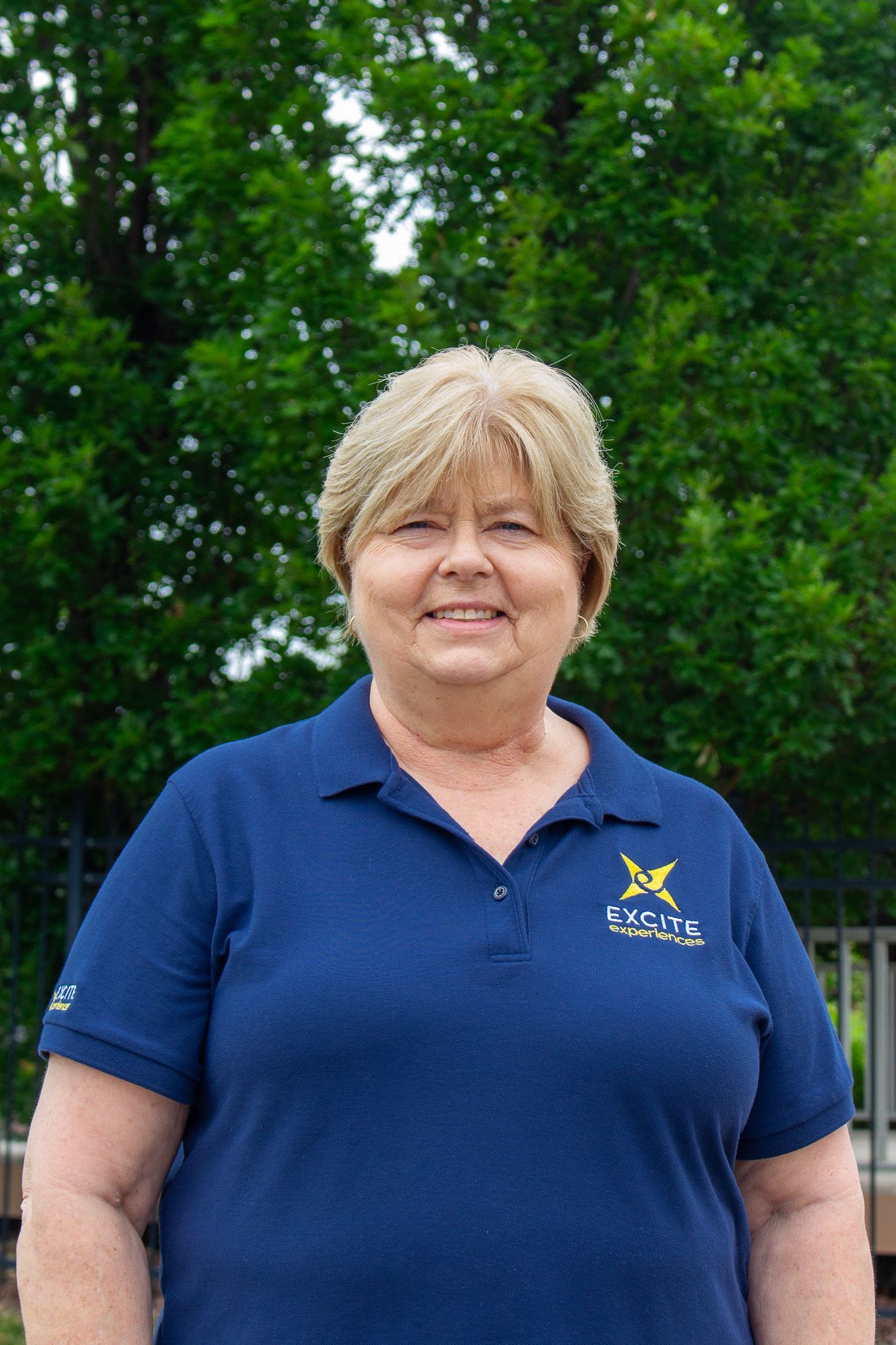 Woman in a blue polo shirt smiles outside with greenery in the background.