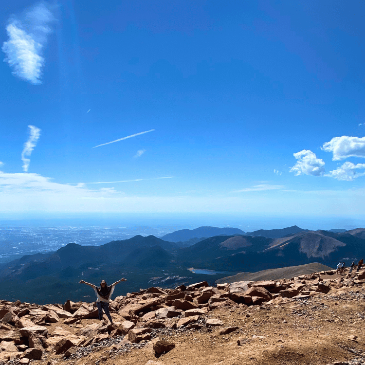 Person jumping joyfully on mountain top, blue sky with clouds, rocky terrain, distant mountains and city view.