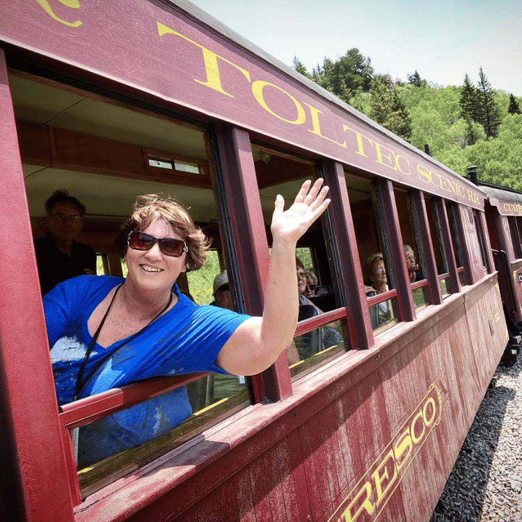 Woman waves from a maroon train car, scenic mountain backdrop.