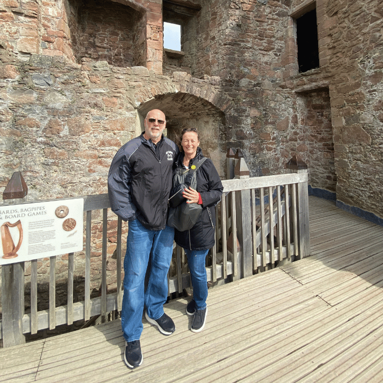 Couple poses on a wooden platform in a stone castle. Man in black jacket and jeans, woman in black jacket.