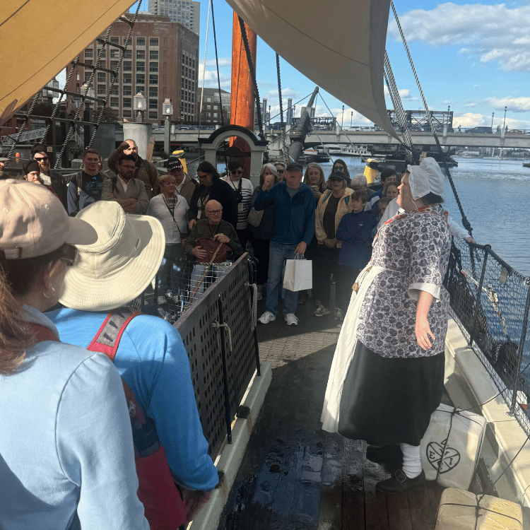 A group of people on a ship listen to a person in historical clothing near a river in Boston.