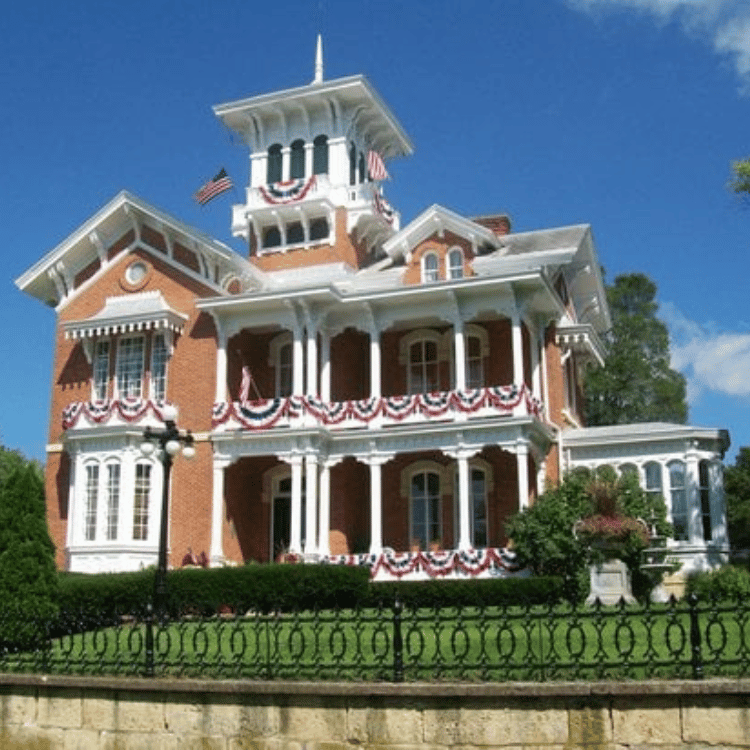 Brick Victorian house with white trim, balconies, and a tower, decorated with red, white, and blue bunting.