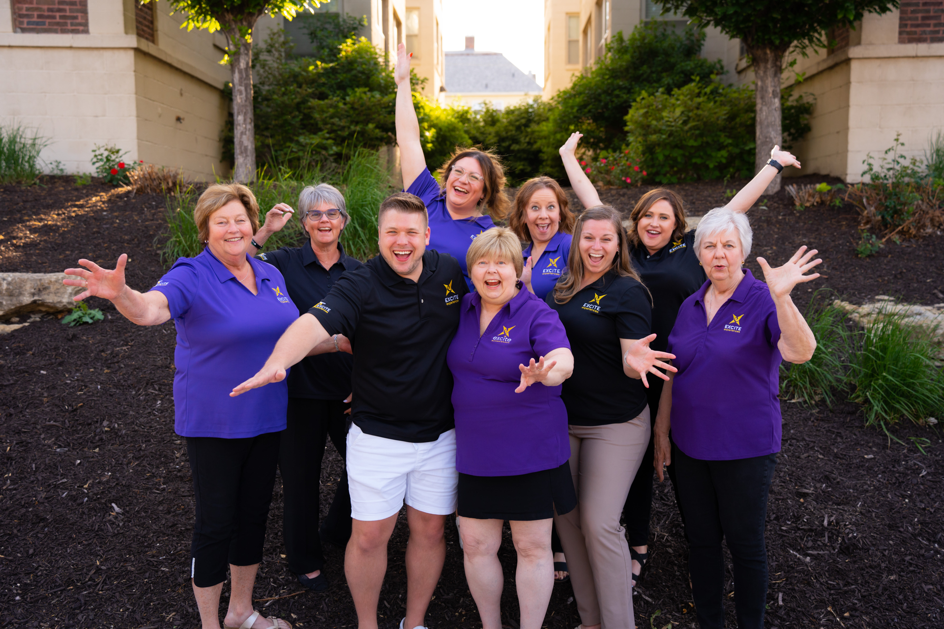 Group of people in purple shirts and black shirts, smiling and raising their arms outside.