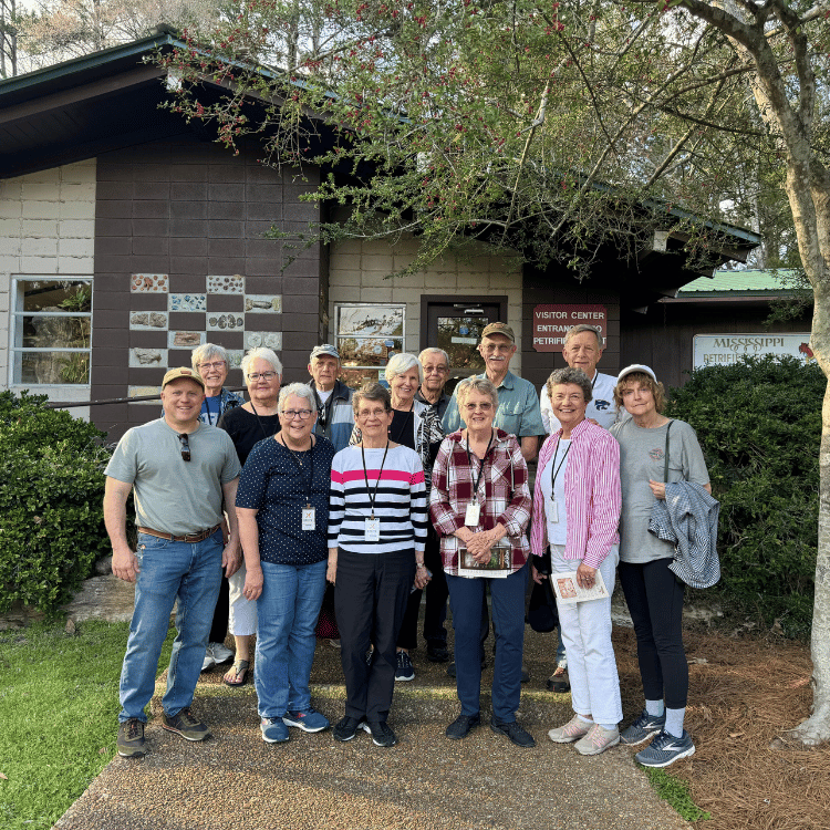 Group of older adults standing outside a building with license plates, smiling in front of a green-roofed building.