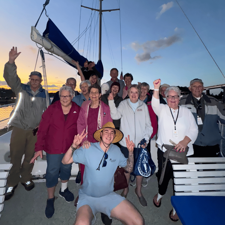 Group of seniors on a boat at dusk, waving and smiling. Young man in front makes peace signs.