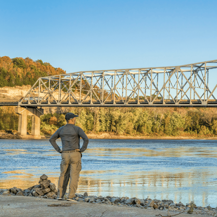 Man standing by a river, looking at a bridge. Blue water, autumn trees, and clear sky.