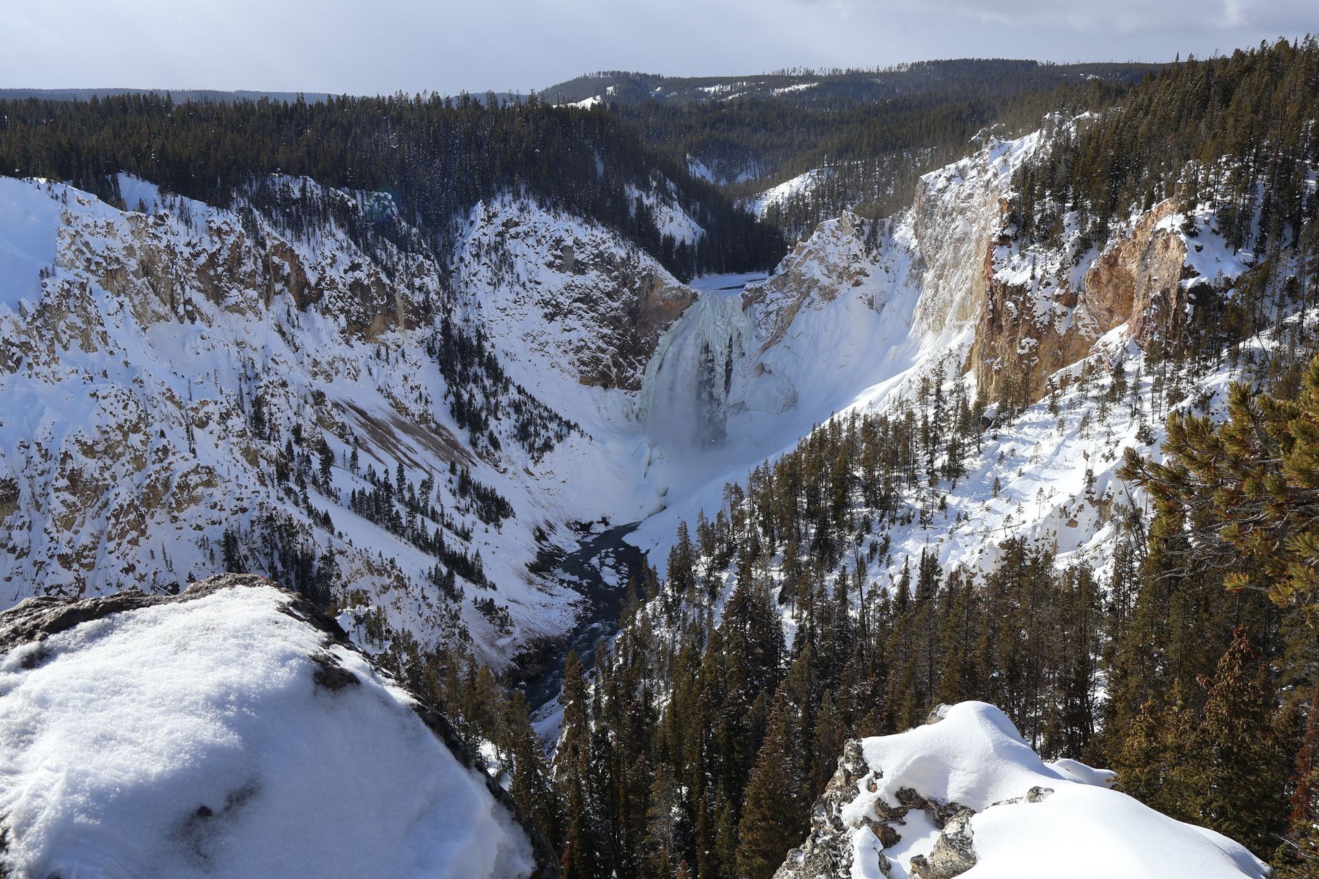 Snow-covered canyon landscape with a waterfall cascading into a deep valley. Evergreen trees and rocky cliffs.