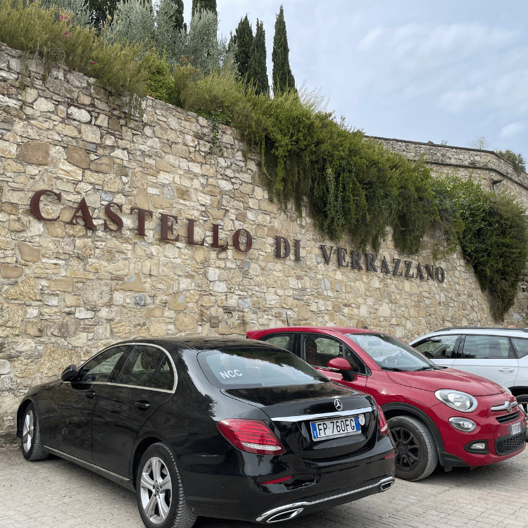 Cars parked in front of Castello di Verrazzano stone wall with signage and greenery.