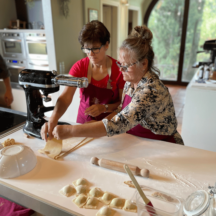 Two people making pasta in a kitchen, using a pasta machine and rolling pin.