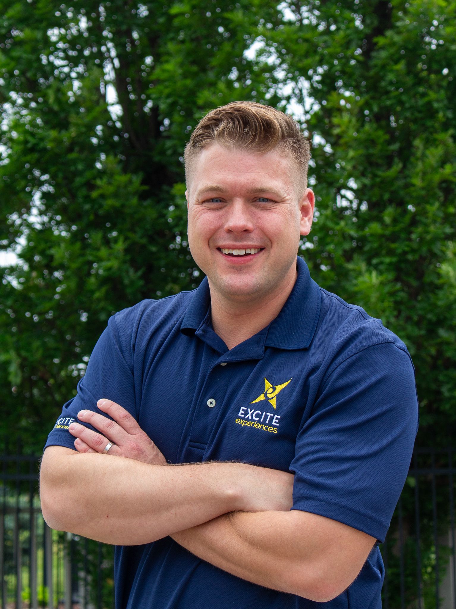Man in blue polo shirt with arms crossed, smiling. Set against a green, leafy background.