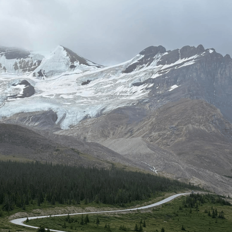 Winding road through a mountain landscape with glacier, forest, and overcast sky.