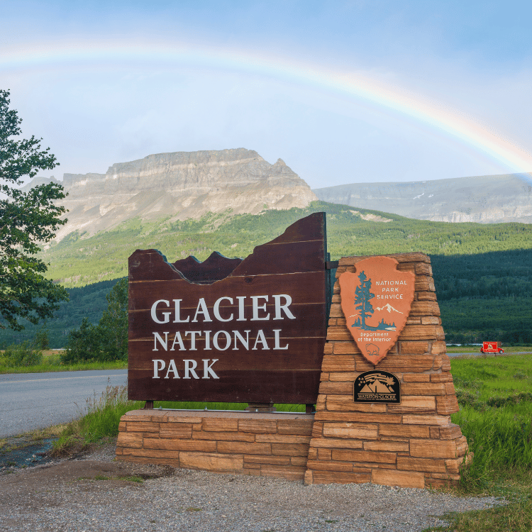 Glacier National Park sign with a mountain backdrop and rainbow in the sky.