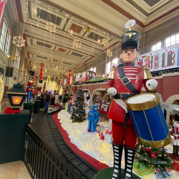 Giant toy soldier stands near a train display decorated for Christmas in a festive building.