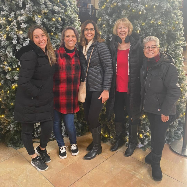 Five women smiling, posing in front of snow-covered Christmas trees.
