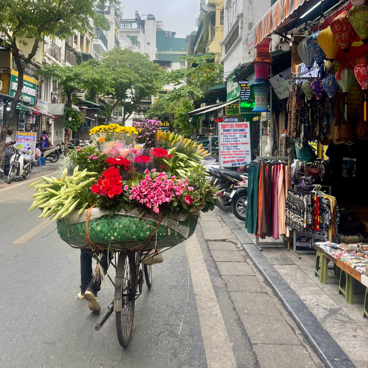 A person rides a bicycle carrying a large basket of colorful flowers on a street lined with shops.