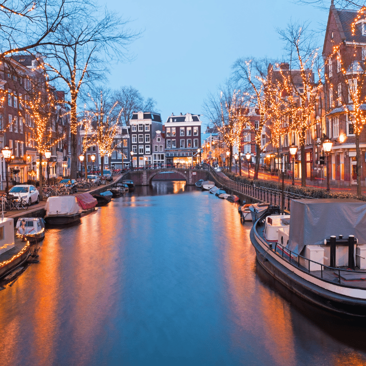 Canal in Amsterdam at dusk, lined with boats, buildings, and festive lights reflecting on the water.