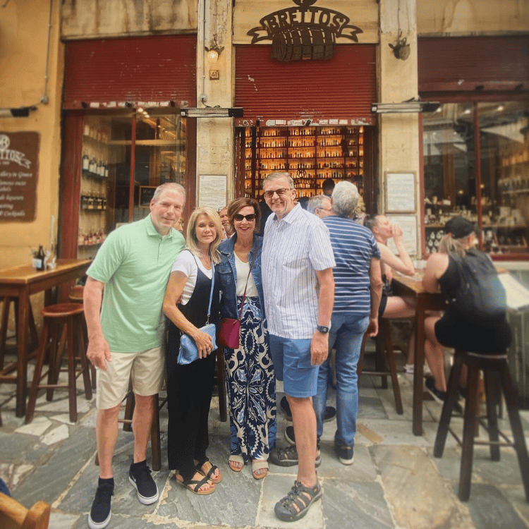 Group of friends outside a shop in Greece. People are standing, smiling, or sitting. Bright sunlight.