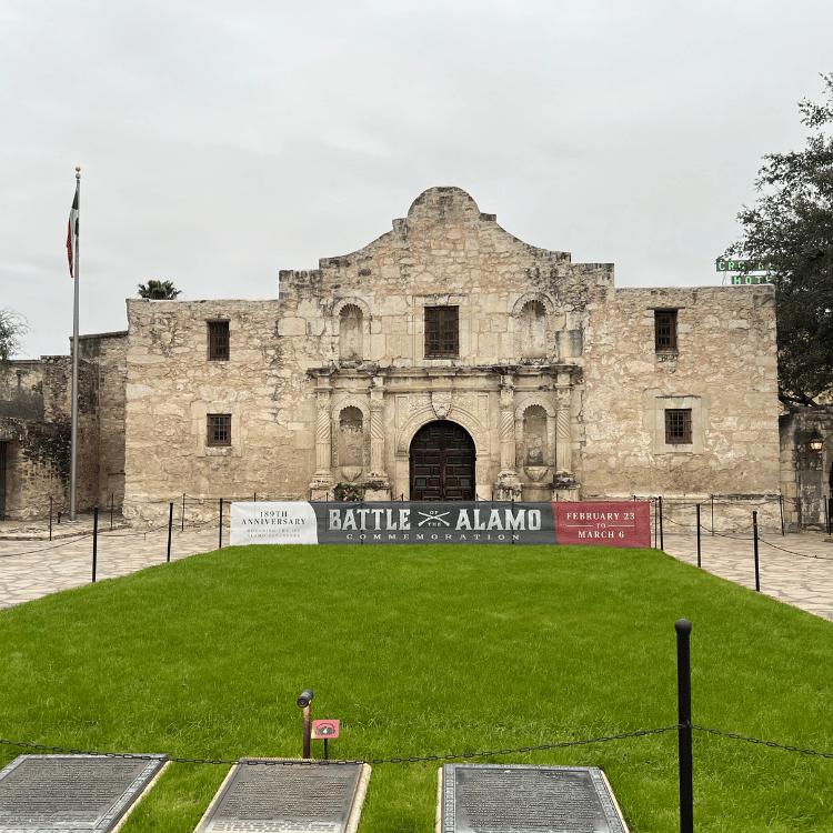 The Alamo Mission, San Antonio, Texas, with a green lawn, a banner, and a cloudy sky.