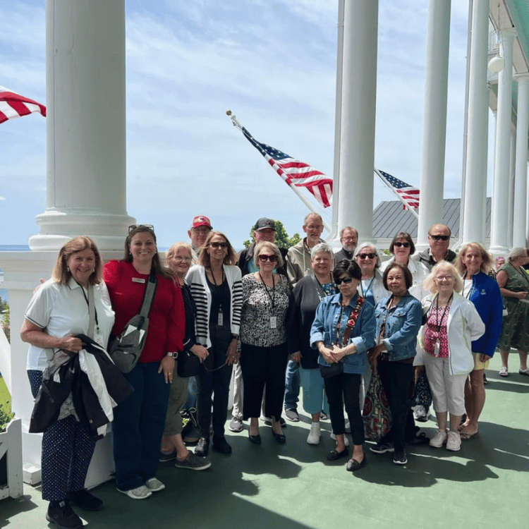 Group of people on a porch with American flags, posing for a photo. Sunny day, white columns, and ocean view.