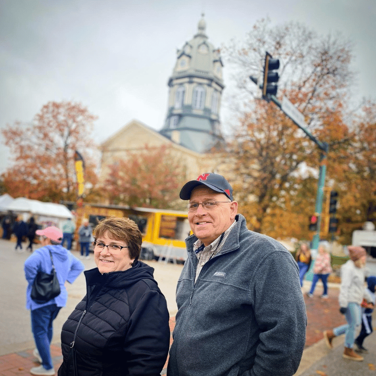 An elderly couple smiles at the camera on a street with a large domed building in the background.