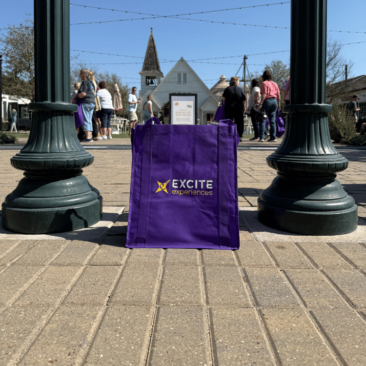 Purple tote bag with "EXCITE" logo on a brick walkway in front of a church, people gathered.