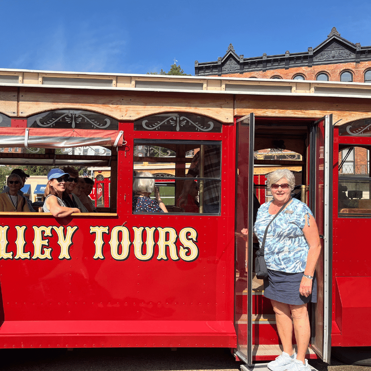 Woman stands in front of red "Valley Tours" trolley. Passengers inside; historic building in background.