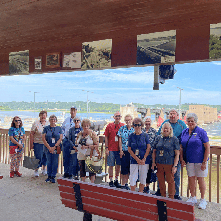 Group of people standing on a deck overlooking a river, under a roof with historical photos.