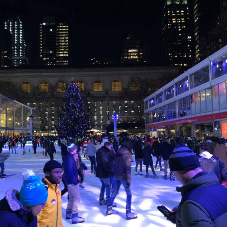 Ice skaters on an outdoor rink at night, lit by blue light, with a Christmas tree and city buildings in the background.