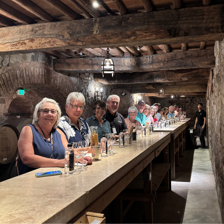 Group of people at a wine tasting in a stone cellar. They are seated at a long wooden table.