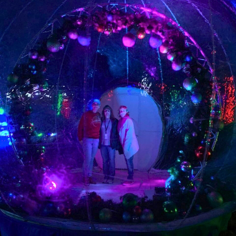 Three people stand inside a brightly lit holiday-themed bubble, surrounded by decorations and colored lights.