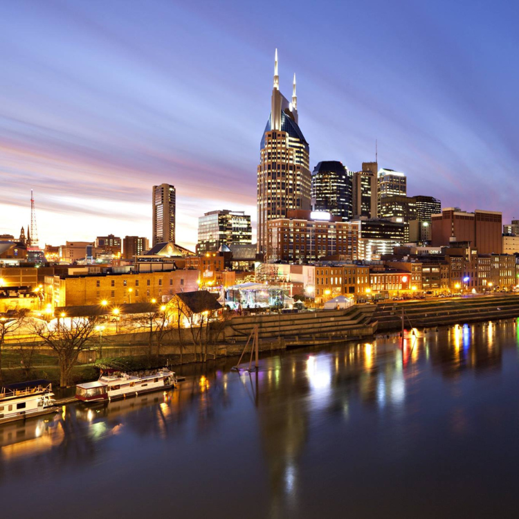 Nashville skyline at dusk reflected in the water with lit buildings, boats, and a colorful sky.