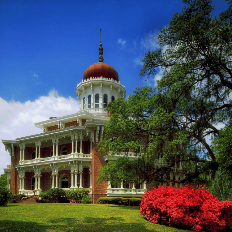Ornate white mansion with a red dome, green lawn, and blue sky. Red azalea bush in the foreground.