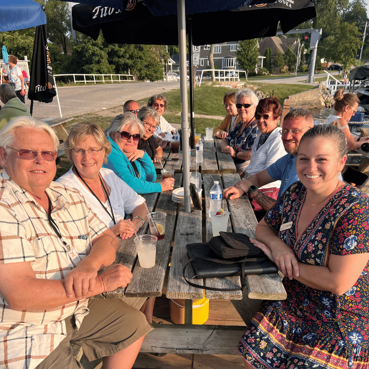 Group of people seated at picnic tables by water, smiling and interacting.