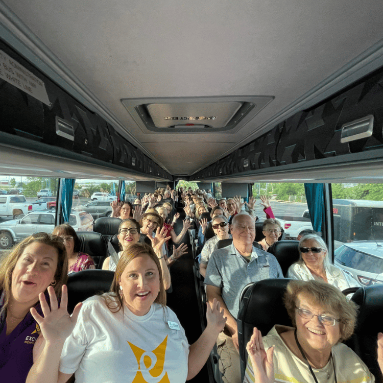 People waving from inside a tour bus, with sunny outdoor view.