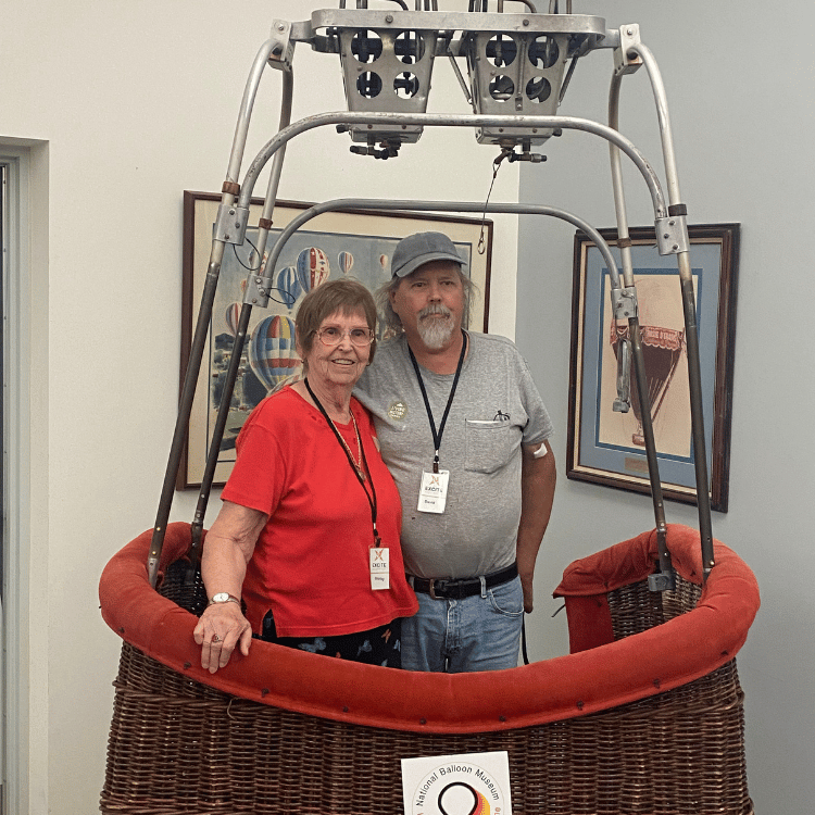 A senior couple poses inside a hot air balloon basket; man in denim, woman in red shirt, indoors.