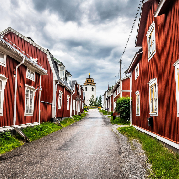Red wooden buildings line a narrow street, leading to a white tower under a cloudy sky.