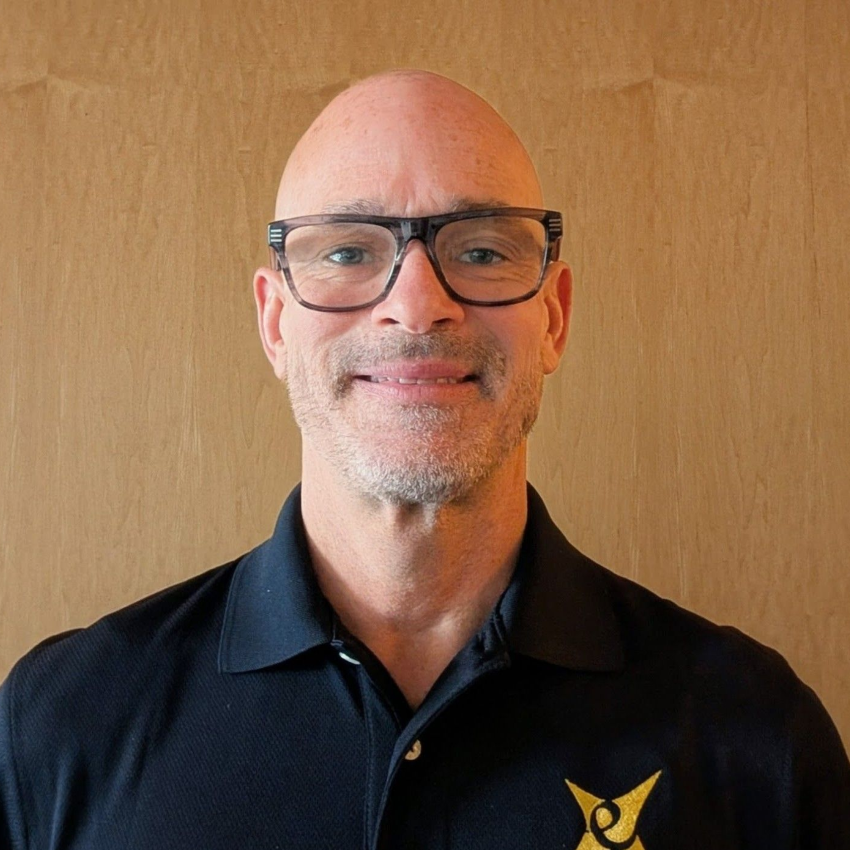 Man with glasses and shaved head smiles, wearing a black polo shirt, against a wood-grain background.