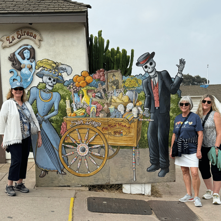 Four women pose near a colorful Día de los Muertos-themed cutout in front of a shop named "La Sirena."